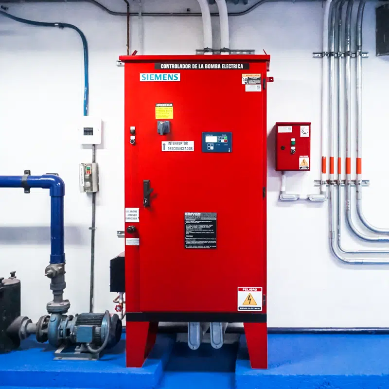 Red Siemens electrical pump control cabinet in a mechanical room with blue floor, connected piping and wiring on the wall behind it.