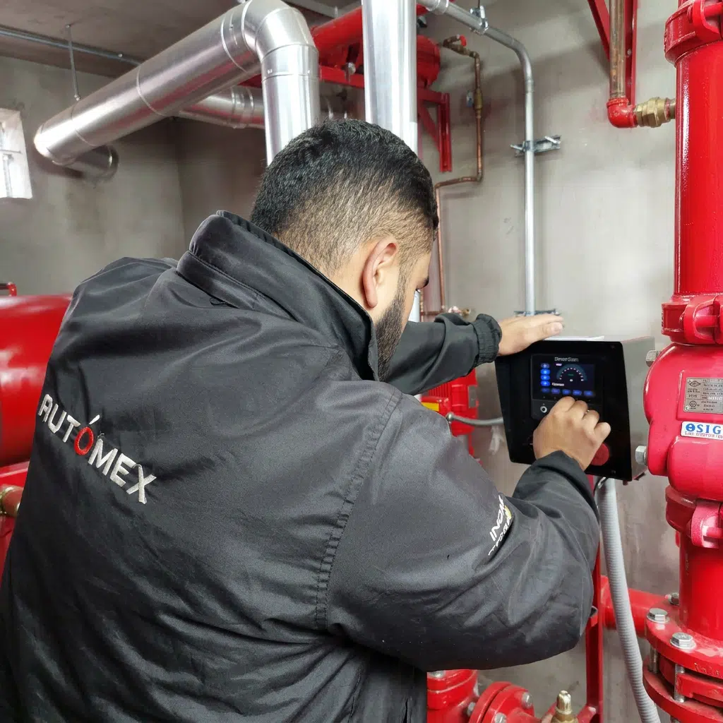 Technician in a black Automex jacket operating a touchscreen control panel among red industrial pipes and valves.