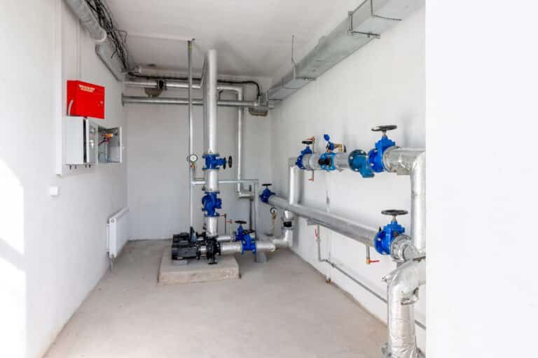 Industrial mechanical room showing insulated silver pipes with blue valve wheels, a black pump on the floor, and a red fire safety cabinet mounted on the left wall.